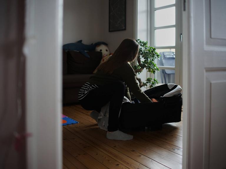 Eine Frau in einer Altbauwohnung beugt sich über eine Babytragetasche.