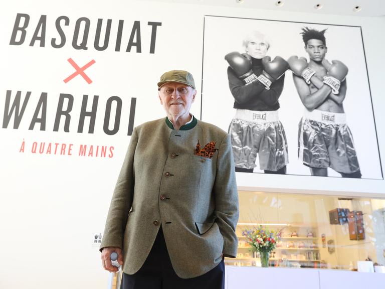 Bruno Bischofberger posiert im Trachtenjanker vor einem Plakat für die Ausstellung "Basquiat X Warhol" in Paris im Jahr 2023