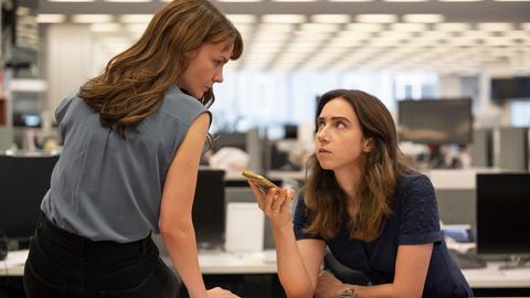 Megan Twohey (Carey Mulligan) und Jodi Kantor (Zoe Kazan, v.l.) im Film She Said von Maria Schrader. Die Figur Megan Twohey sitzt auf dem Schreibtisch, Jodi Kantor lehnt daran. Sie hält ein Mobiltelefon. Beide schauen einander an. Im Hintergrund ist unscharf ein Großraumbüro zu sehen. 