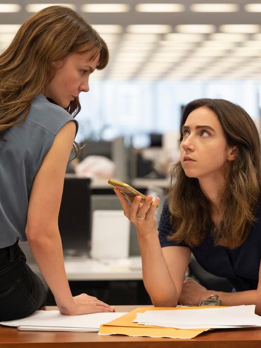 Megan Twohey (Carey Mulligan) und Jodi Kantor (Zoe Kazan, v.l.) im Film She Said von Maria Schrader. Die Figur Megan Twohey sitzt auf dem Schreibtisch, Jodi Kantor lehnt daran. Sie hält ein Mobiltelefon. Beide schauen einander an. Im Hintergrund ist unscharf ein Großraumbüro zu sehen. 