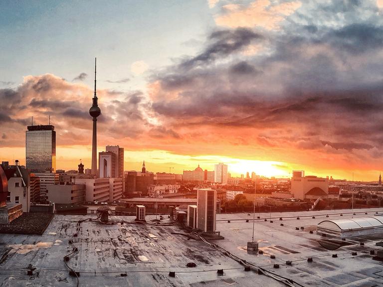 Blick über Berlin im Sonnenuntergang von einem betonierten Dach aus. Der Fernsehturm zeichnet sich deutlich vor dem aufgeregten Himmel ab.