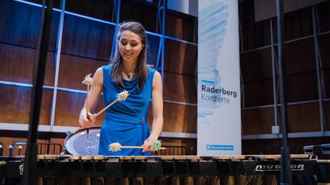 Eine junge Frau in blauem Kleid spielt Schlagwerk auf einer Bühne.