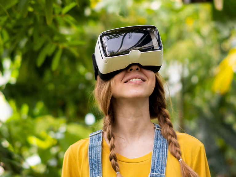 Eine Frau trägt in grüner Umgebung eine Virtual Reality (VR) Brille und schaut erfreut in den Himmel.