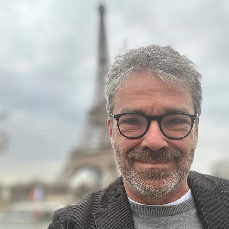 Ein Mann mit kurzen grauen Haaren und Brille lächelt in die Kamera, im Hintergrund erkennt man den Pariser Eiffelturm