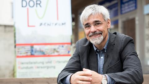 Ein Mann mit grauen Haaren, Brille, Schnauzer und Kinnbart sitzt mit gefalteten Händen vor dem Schild eines dorfladens.
