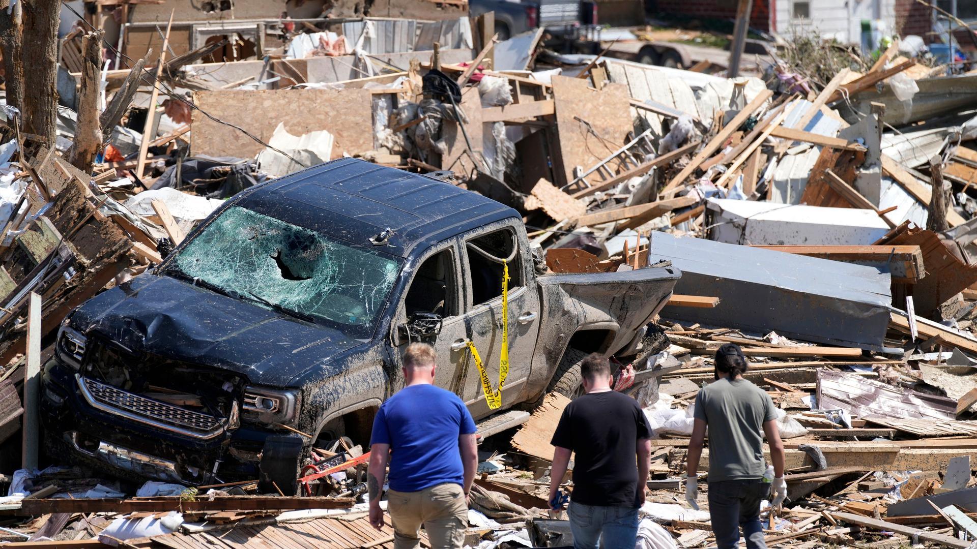 Menschen beschauen sich die Zerstörung durch einen Tornado in Greenfield, Iowa an. Man sieht Trümmer und ein völlig zerstörtes Auto.