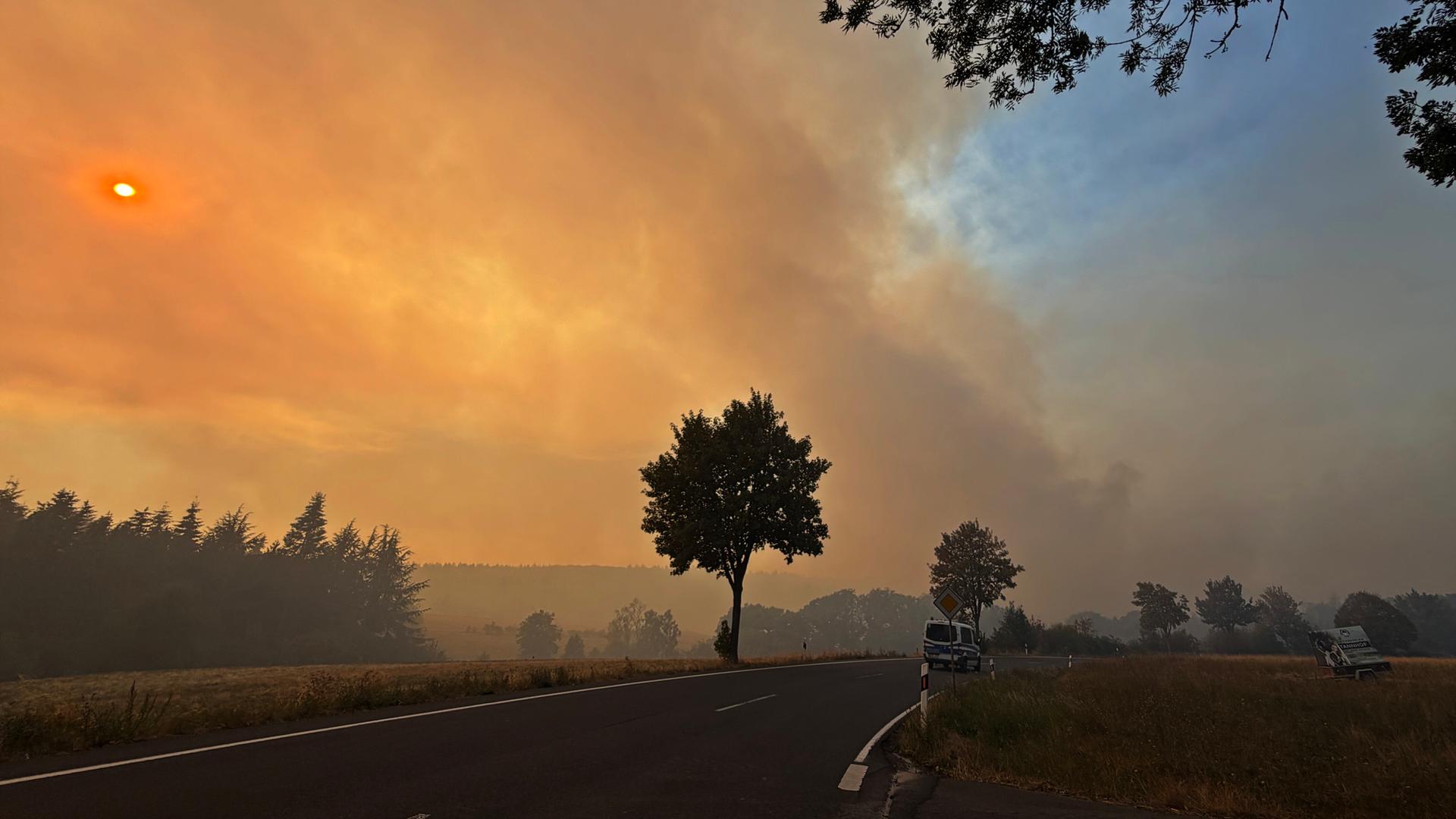 Rauchschwaden und Feuerschein über einer Landstraße, auf der ein Polizeiauto fährt: Waldbrand im Hürtgenwald