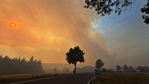 Rauchschwaden und Feuerschein über einer Landstraße, auf der ein Polizeiauto fährt: Waldbrand im Hürtgenwald