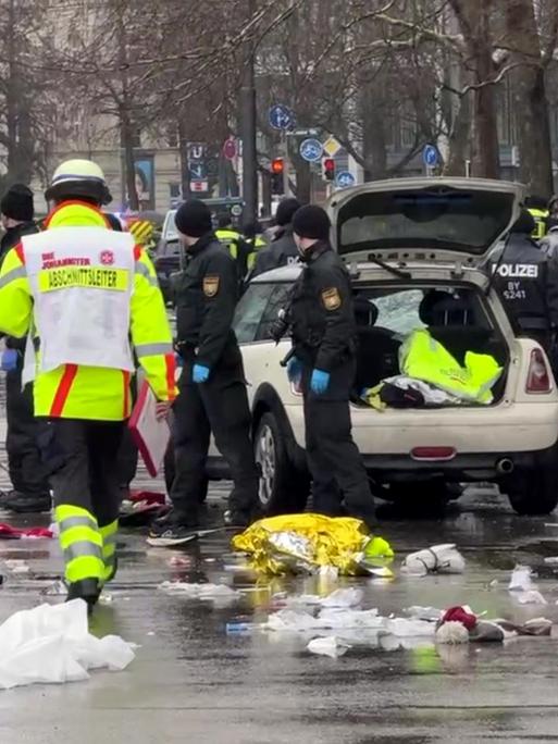 Rettungskräfte stehen am Einsatzort um ein Auto. In der Münchner Innenstadt ist ein Fahrzeug in eine Menschengruppe gefahren. Auf dem Boden liegen medizinische und persönliche Gegenstände.