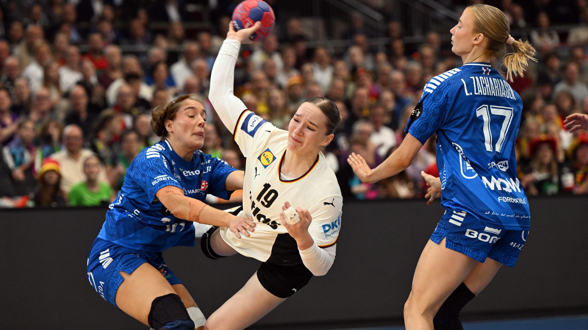 Nieke Kühne setzt sich im deutschen Nationaltrikot mit einem akrobatischen Sprung gegen zwei Gegenspielerinnen durch. Dabei führt sie in ihrer rechen Hand den Ball.