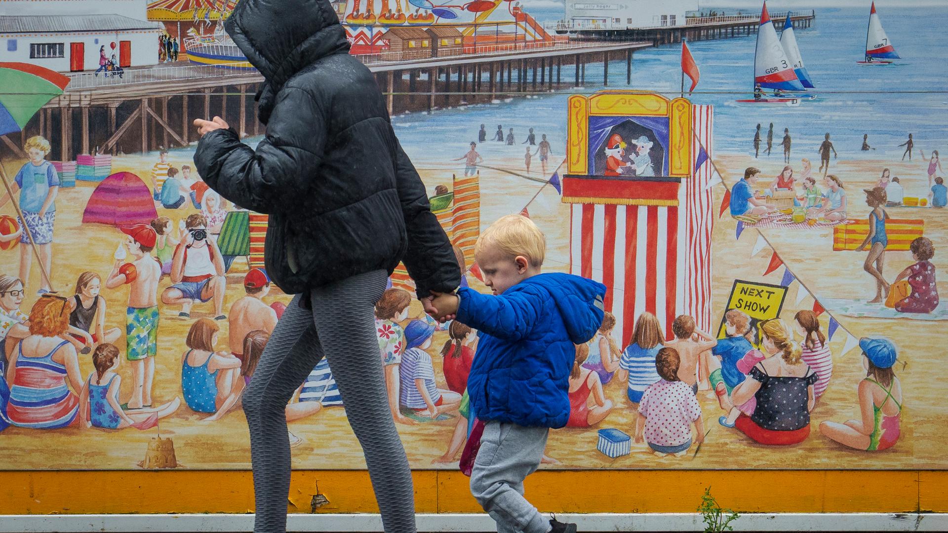 Eine Frau und ein Kind gehen in Clacton-on-Sea spazieren. Im Hintergrund ist ein Cartoon des britischen Seebads zu sehen. 