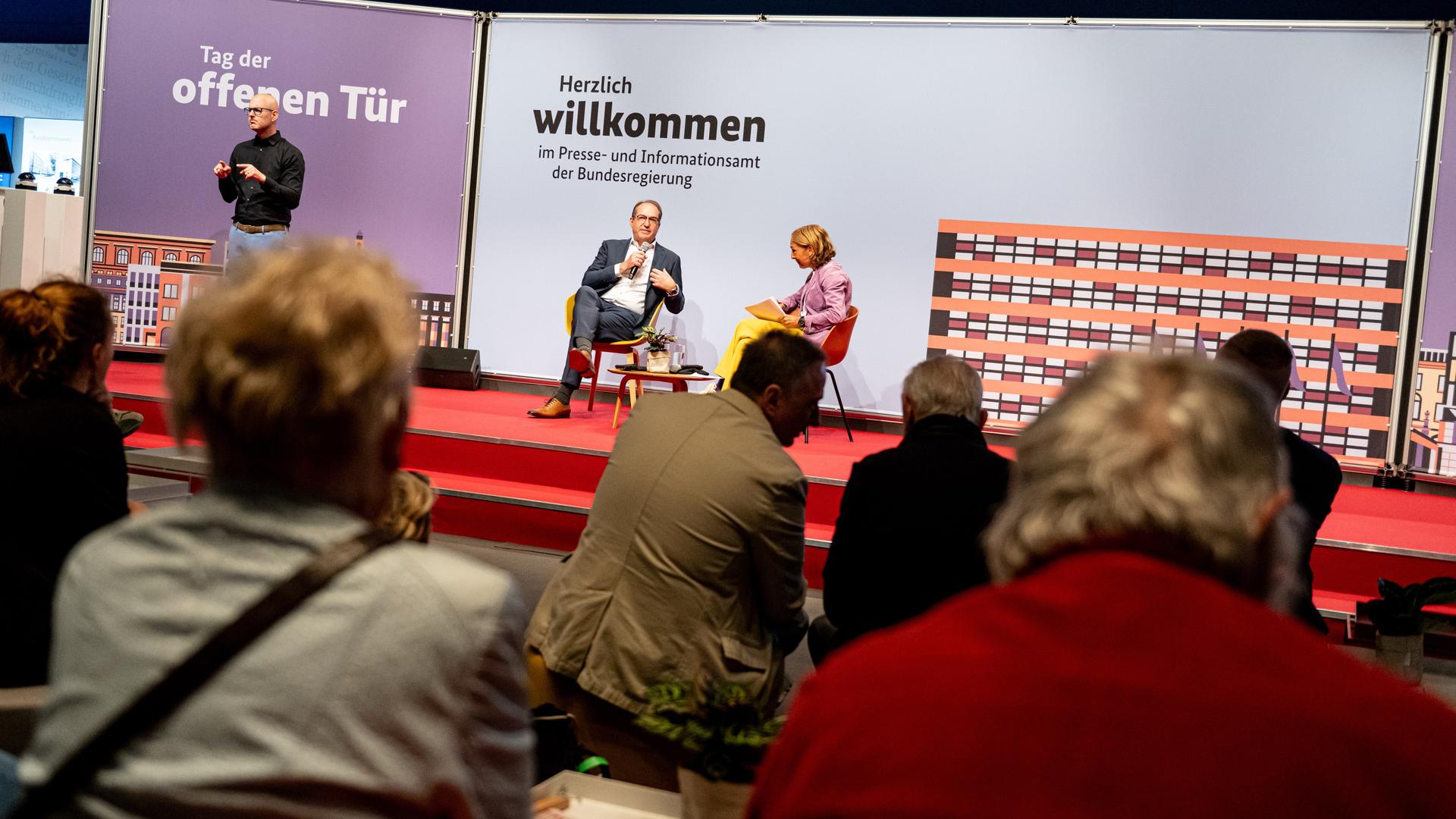 Innenminister Dobrindt sitzt im Bildhintergrund auf einer Bühne mit einem Mikrofon in der Hand. Neben ihm sitzt eine Frau, die ihn befragt. Eine Gruppe von Menschen hört zu. Auf einer Stellwand steht "Tag der offenen Tür" und "Herzlich Willkommen". Innenminister Dobrindt sitzt im Bildhintergrund auf einer Bühne mit einem Mikrofon in der Hand. Neben ihm sitzt eine Frau, die ihn befragt. Eine Gruppe von Menschen hört zu. Auf einer Stellwand steht "Tag der offenen Tür" und "Herzlich Willkommen".