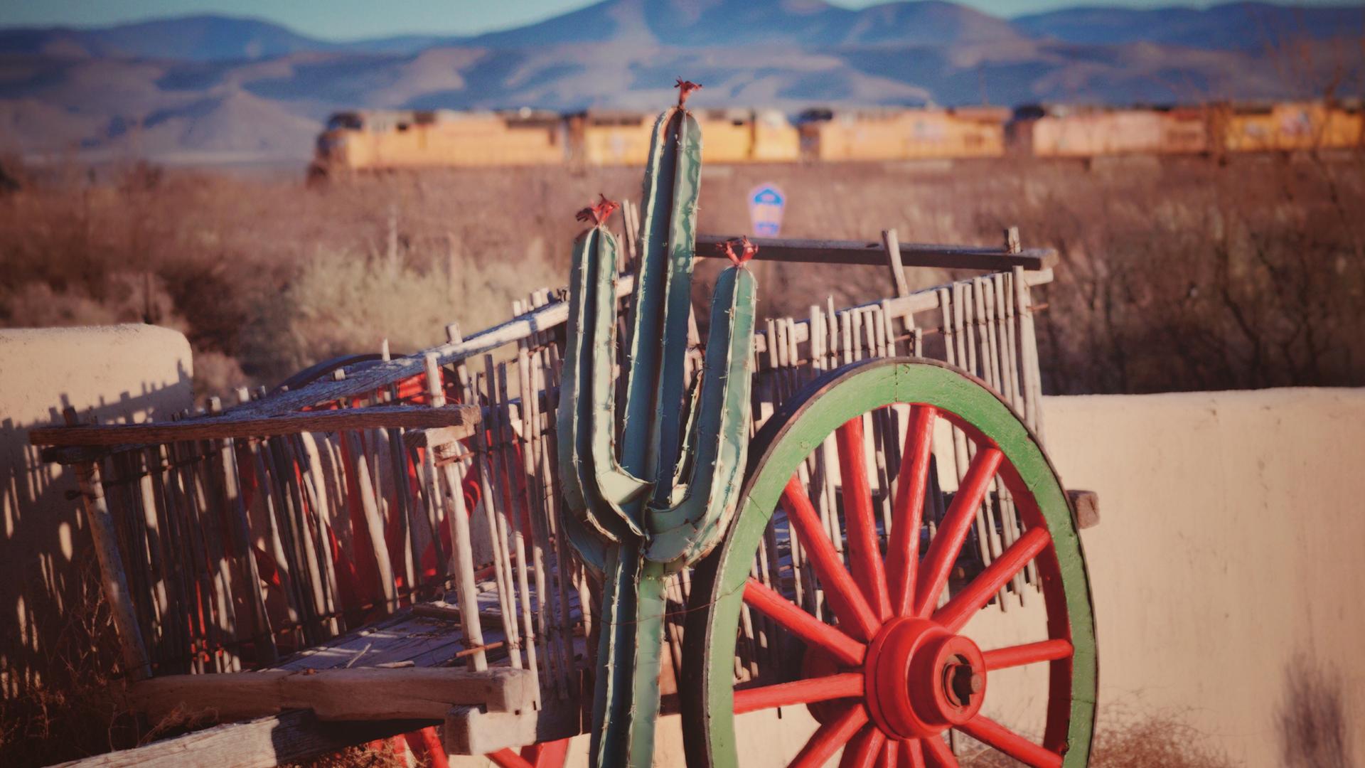 Ein Kaktus vor einem Handkarren aus Holz vor einer Wild-West-Kulisse.