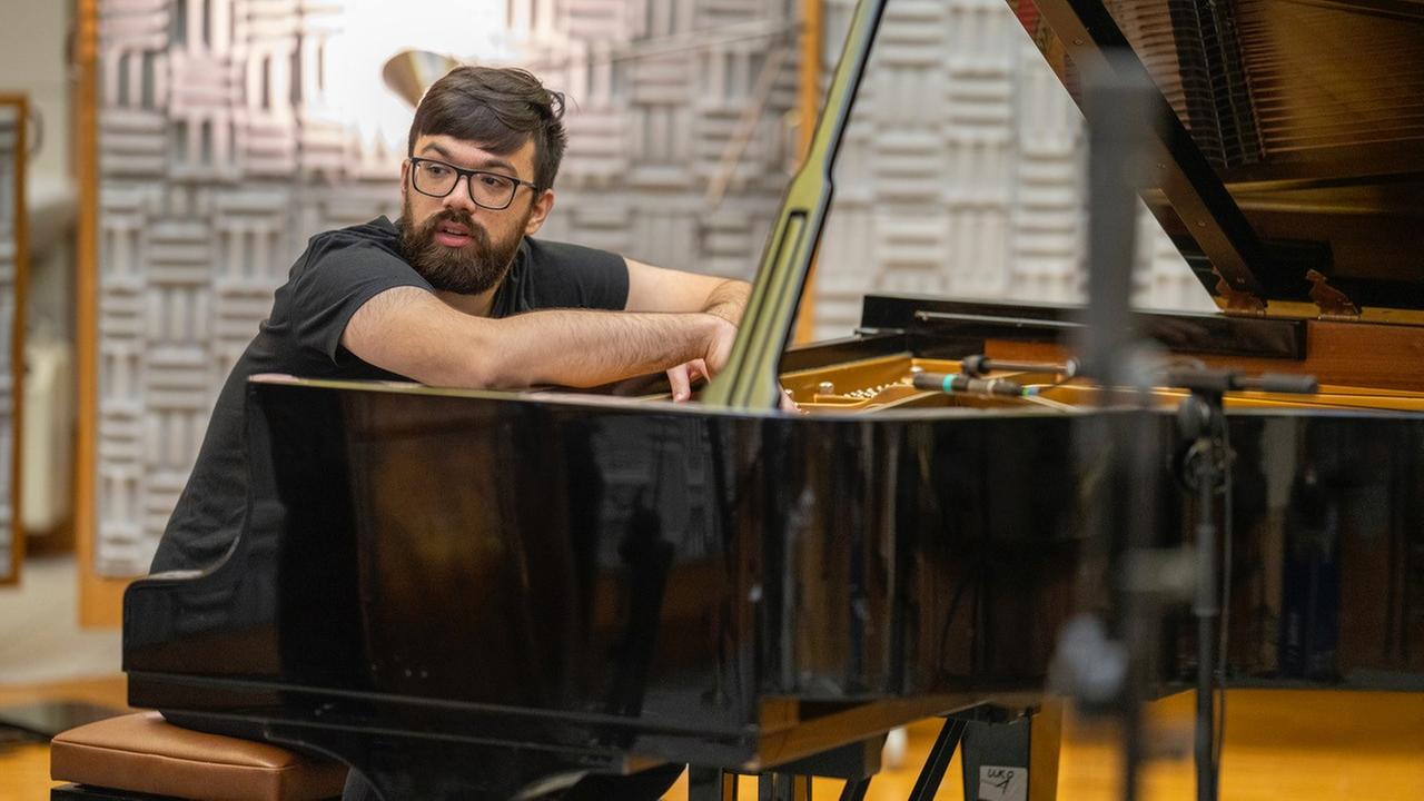 Ein bärtiger Mann mit schwarzer Brille schaut, über seinen Klavierflügel gelehnt, zur rechten Seite.