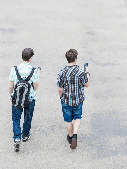 Blick von hinten auf zwei Jugendliche, die auf einer Straße gehen und Smartphones in der Hand halten.