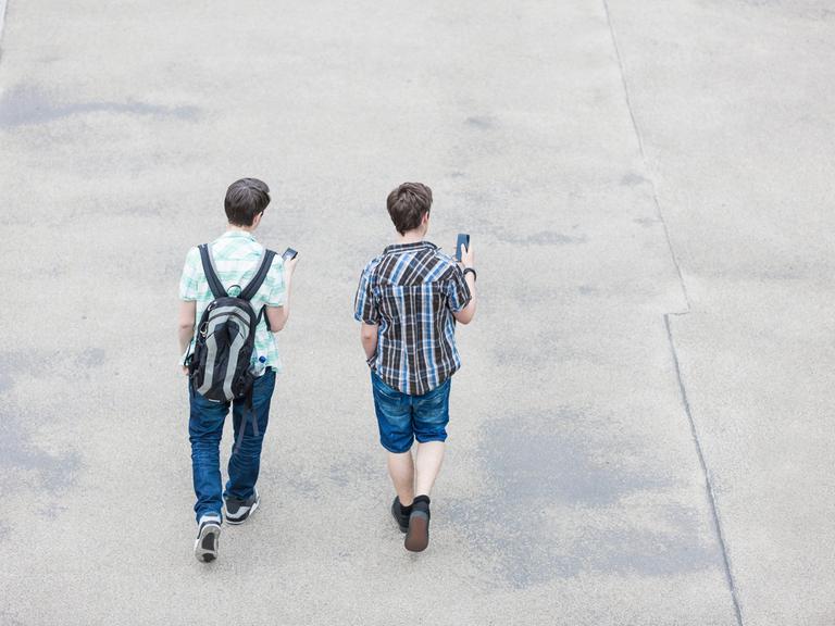 Blick von hinten auf zwei Jugendliche, die auf einer Straße gehen und Smartphones in der Hand halten.