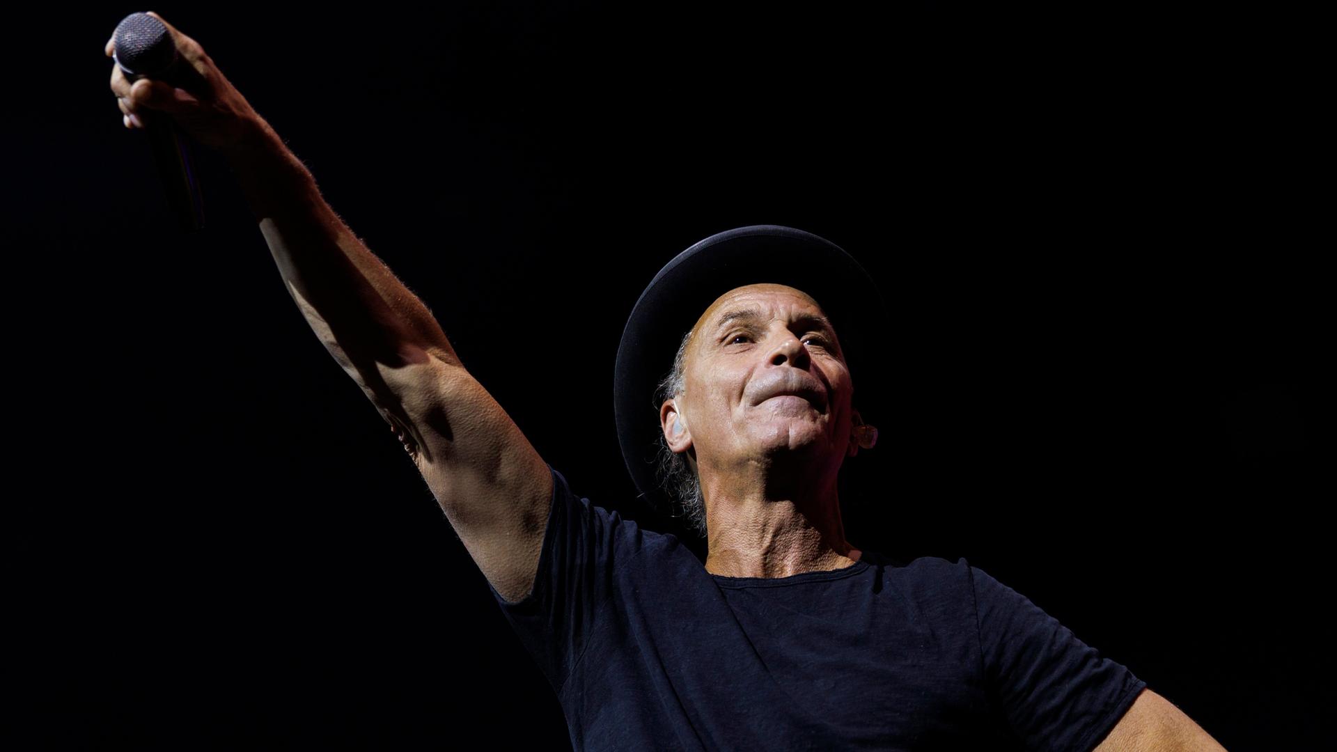 Sänger Peter Brings trägt T-Shirt und Hut und streckt seinen rechten Arm mit dem Mikrofon in die Höhe.