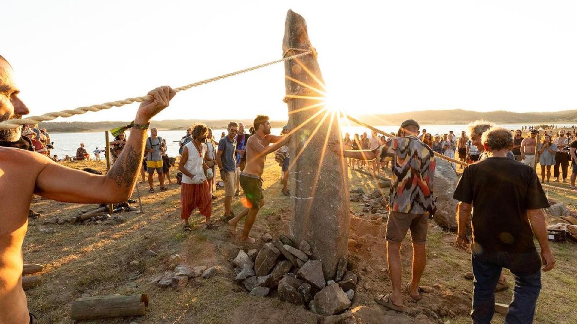 Junge Menschen auf dem Boom-Festival in Portugal richten gemeinsam einen großen Stein auf dem Festivalgelände auf.