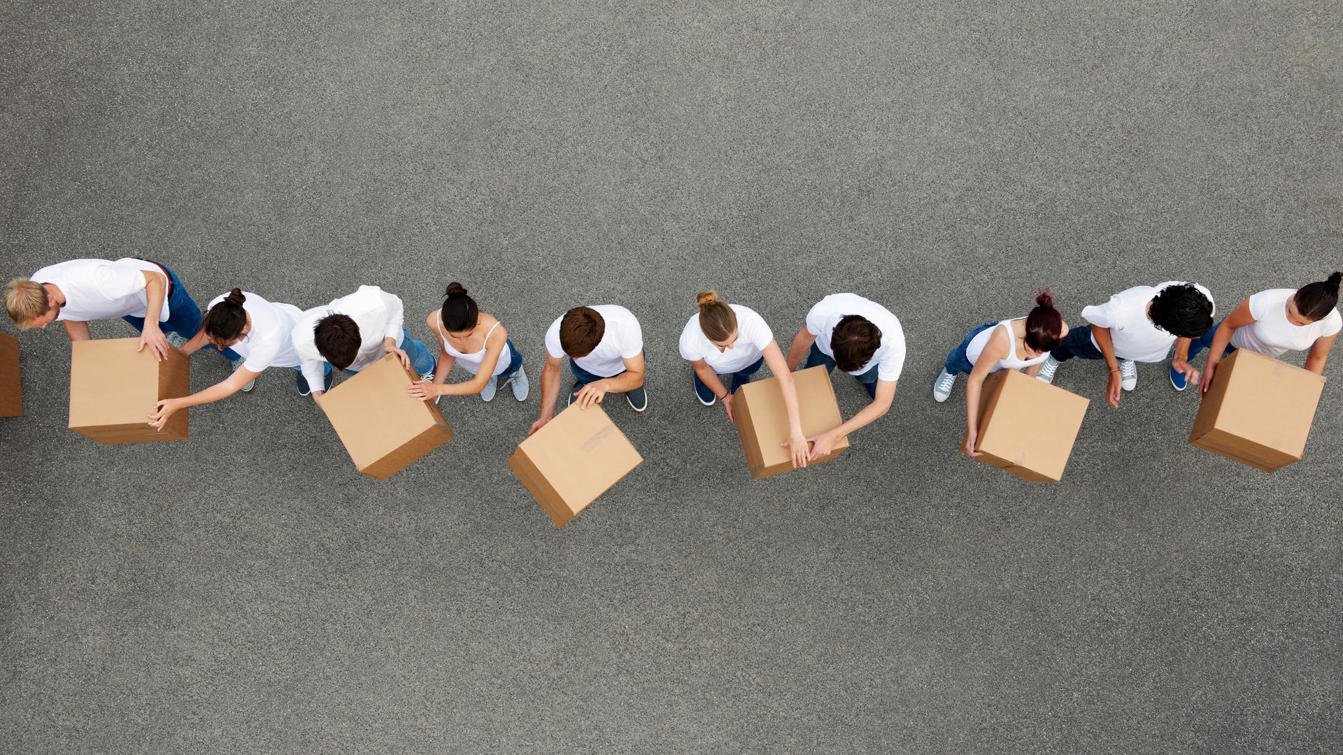 Luftaufnahme von Personen die sich Kartons gegenseitig weiterreichen. Die Personen tragen weiße T-Shirts und stehen auf einem grauen Betongrund. Luftaufnahme von Personen die sich Kartons gegenseitig weiterreichen. Die Personen tragen weiße T-Shirts und stehen auf einem grauen Betongrund.
