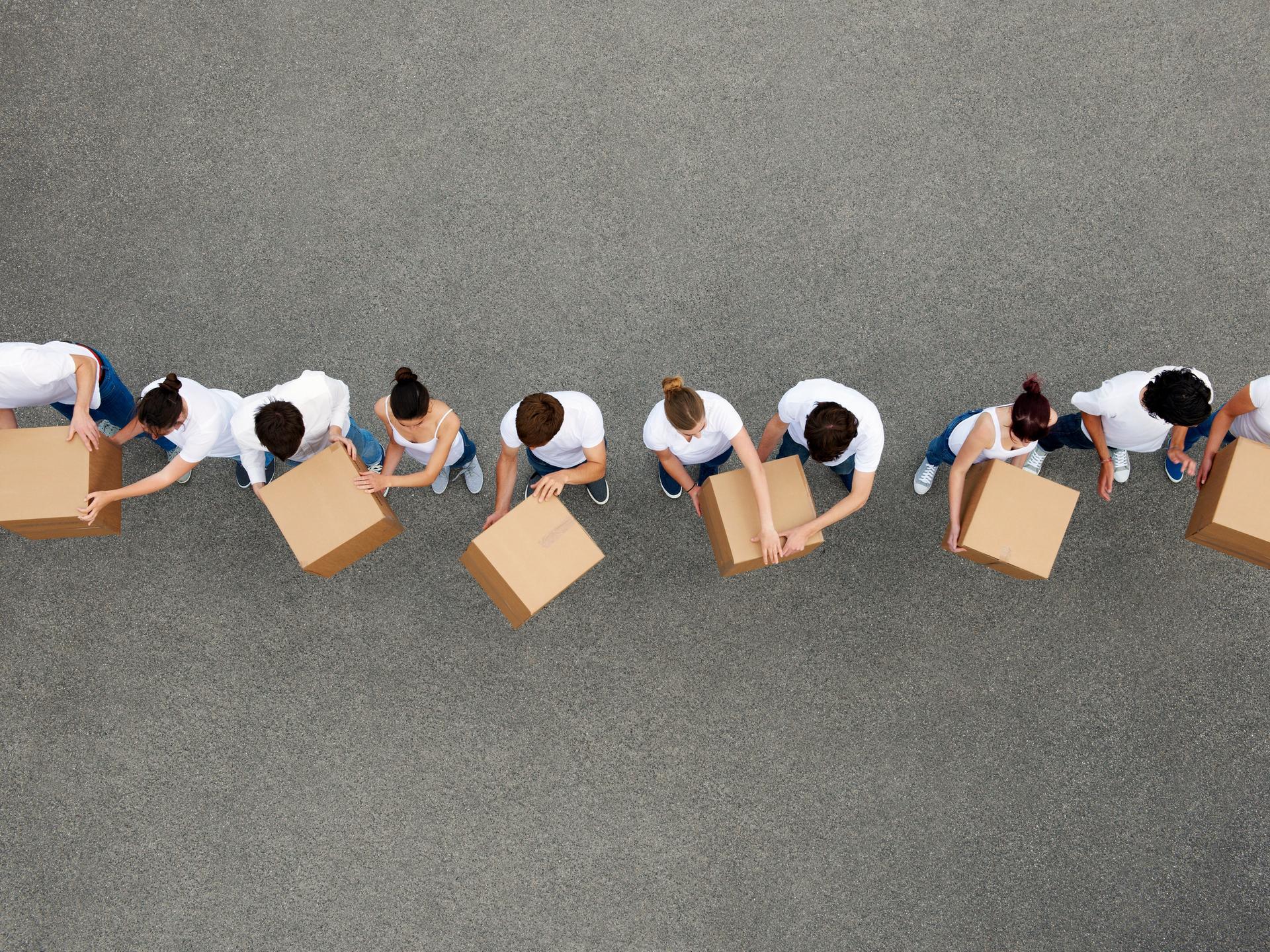 Luftaufnahme von Personen die sich Kartons gegenseitig weiterreichen. Die Personen tragen weiße T-Shirts und stehen auf einem grauen Betongrund.