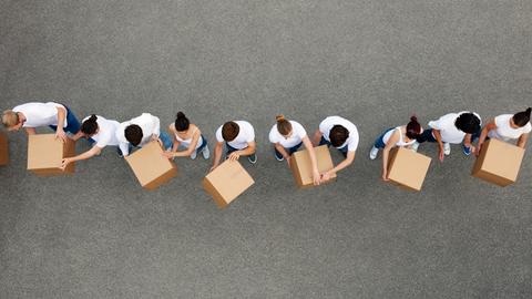 Luftaufnahme von Personen die sich Kartons gegenseitig weiterreichen. Die Personen tragen weiße T-Shirts und stehen auf einem grauen Betongrund. Luftaufnahme von Personen die sich Kartons gegenseitig weiterreichen. Die Personen tragen weiße T-Shirts und stehen auf einem grauen Betongrund.