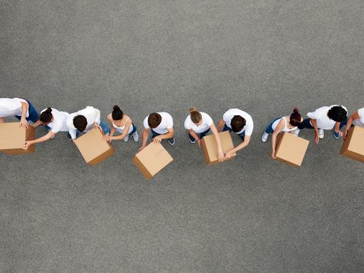 Luftaufnahme von Personen die sich Kartons gegenseitig weiterreichen. Die Personen tragen weiße T-Shirts und stehen auf einem grauen Betongrund.