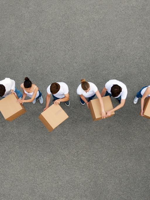 Luftaufnahme von Personen die sich Kartons gegenseitig weiterreichen. Die Personen tragen weiße T-Shirts und stehen auf einem grauen Betongrund.