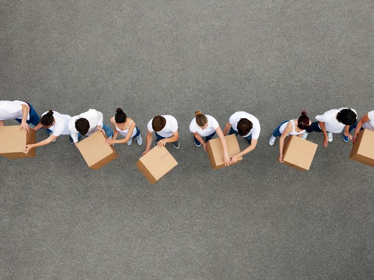 Luftaufnahme von Personen die sich Kartons gegenseitig weiterreichen. Die Personen tragen weiße T-Shirts und stehen auf einem grauen Betongrund.