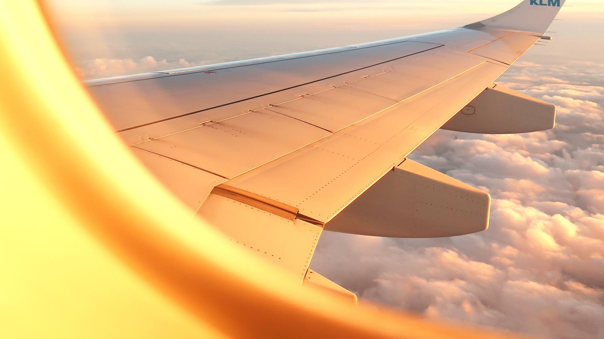Blick aus dem Kabinenfenster auf die Tragfläche eines Flugzeuges. Im oberen Bereich blauer Himmel, darunter eine dichte, weiße Wolkendecke.