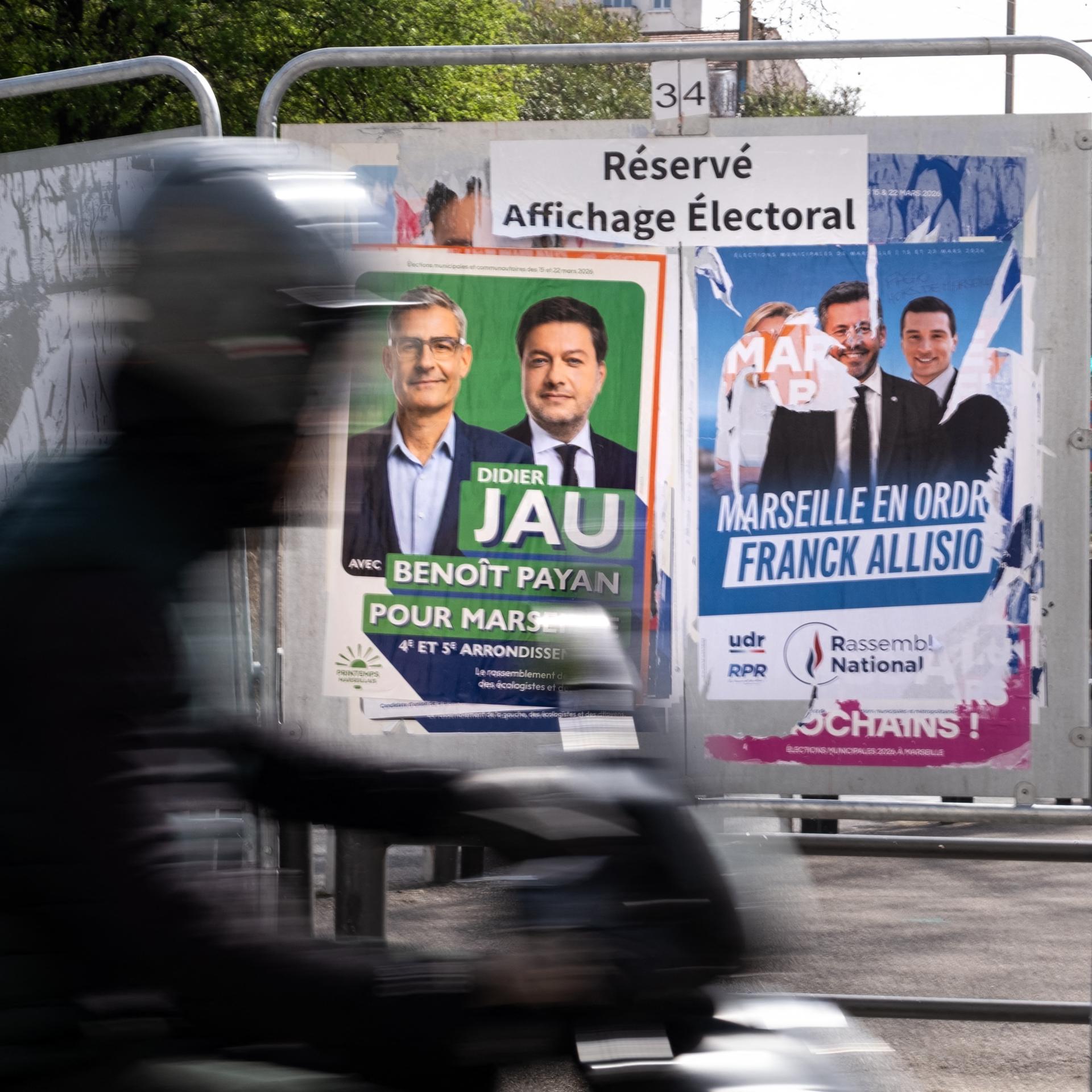 Zu sehen sind Wahlplakate der Bürgermeisterkandidaten von Marseille. Im Vordergrund fährt eine Person auf dem Motorrad vorbei. 
