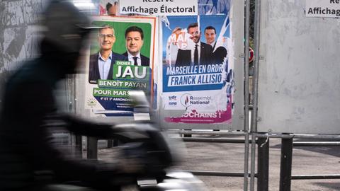 Zu sehen sind Wahlplakate der Bürgermeisterkandidaten von Marseille. Im Vordergrund fährt eine Person auf dem Motorrad vorbei. 