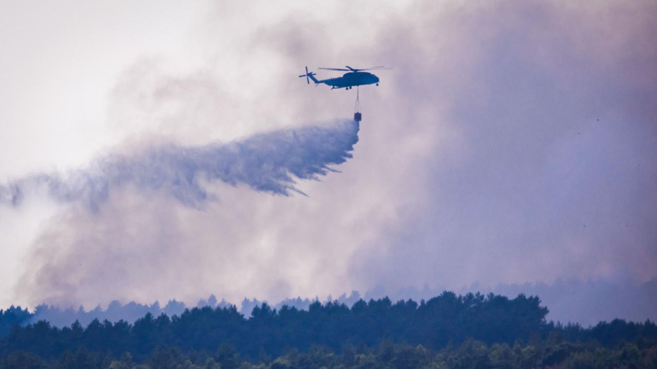 Ein Hubschrauber fliegt über einem bewaldeten Gebiet und lässt aus einem Behälter eine große Menge Löschwasser fallen. Dichter grauer Rauch liegt über den Baumkronen und verdeckt weite Teile des Himmels. Ein Hubschrauber fliegt über einem bewaldeten Gebiet und lässt aus einem Behälter eine große Menge Löschwasser fallen. Dichter grauer Rauch liegt über den Baumkronen und verdeckt weite Teile des Himmels.
