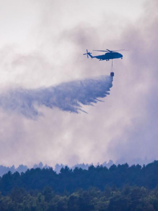 Ein Hubschrauber fliegt über einem bewaldeten Gebiet und lässt aus einem Behälter eine große Menge Löschwasser fallen. Dichter grauer Rauch liegt über den Baumkronen und verdeckt weite Teile des Himmels.