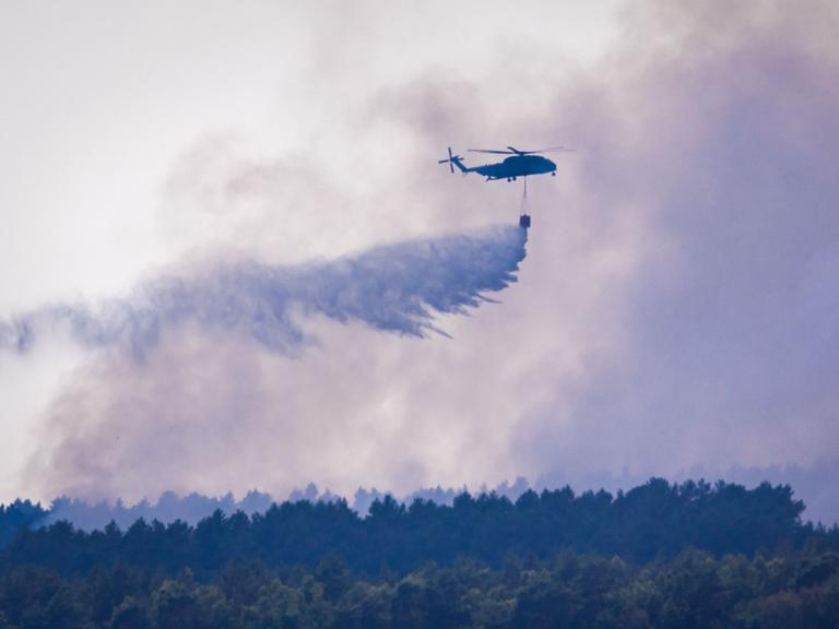 Ein Hubschrauber fliegt über einem bewaldeten Gebiet und lässt aus einem Behälter eine große Menge Löschwasser fallen. Dichter grauer Rauch liegt über den Baumkronen und verdeckt weite Teile des Himmels.