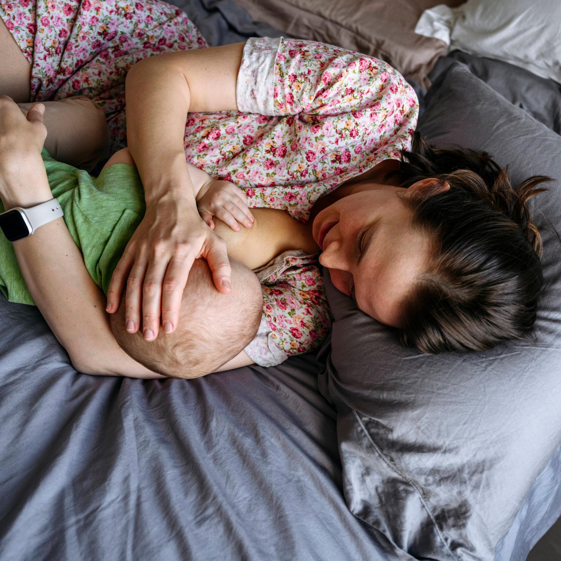 Eine Frau liegt engumschlungen mit einem Baby auf einem Bett und stillt es.