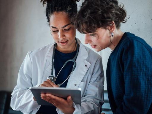Eine Ärztin und eine Patientin schauen gemeinsam auf ein Tablet, das die Ärztin in der Hand hält.