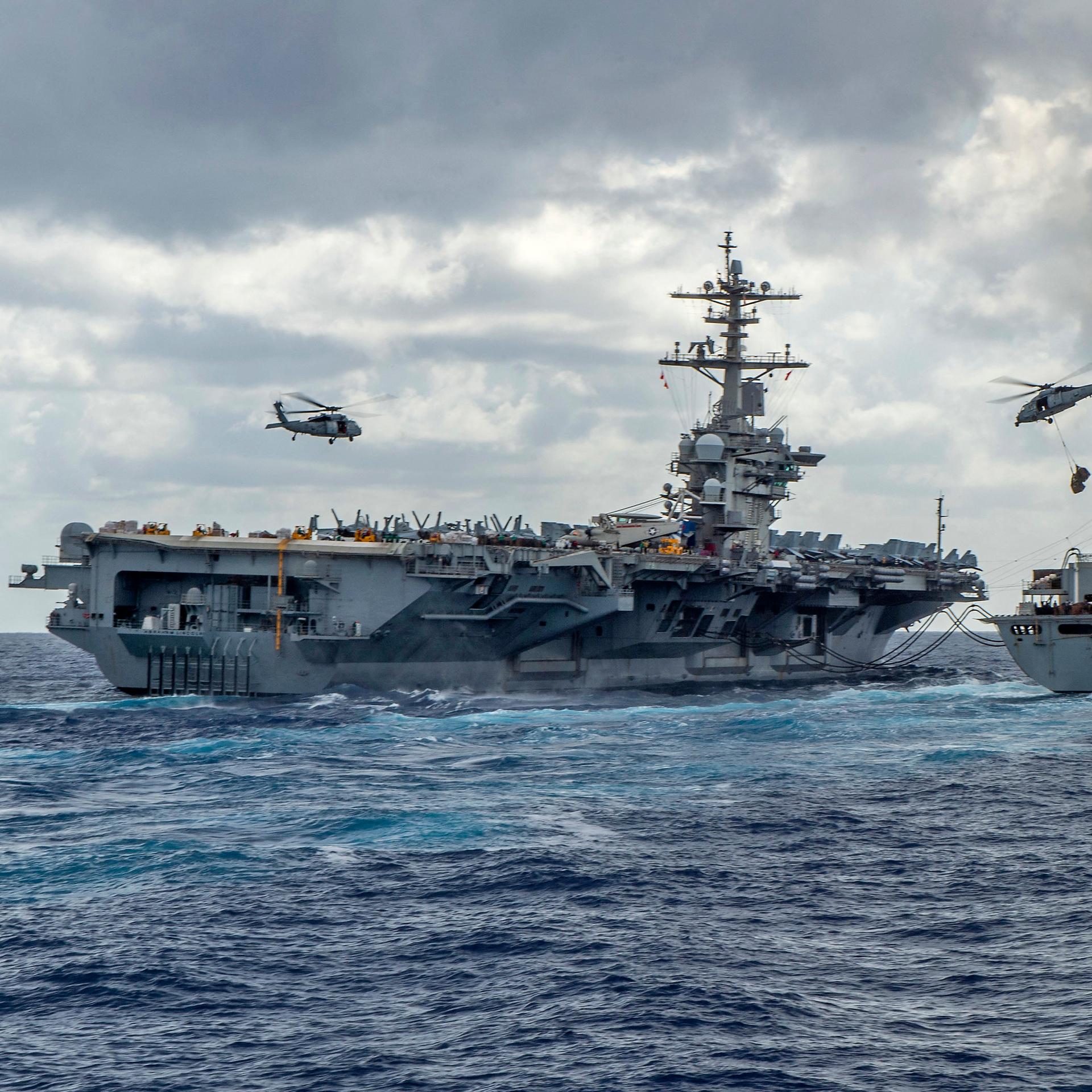 Der US-Flugzeugträger USS Abraham Lincoln mit einem Begleitschiff auf hoher See, darüber fliegen mehrere Hubschrauber