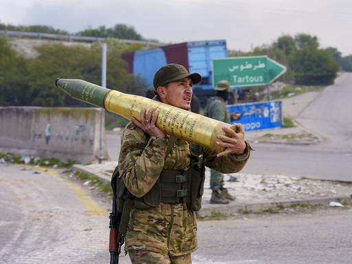 Ein Soldat trägt eine Granate. Er hält sie mit beiden Händen und stützt sie auf seiner rechten Schulter ab. Im Hintergrund ist ein Wegweiser nach Tartous zu sehen