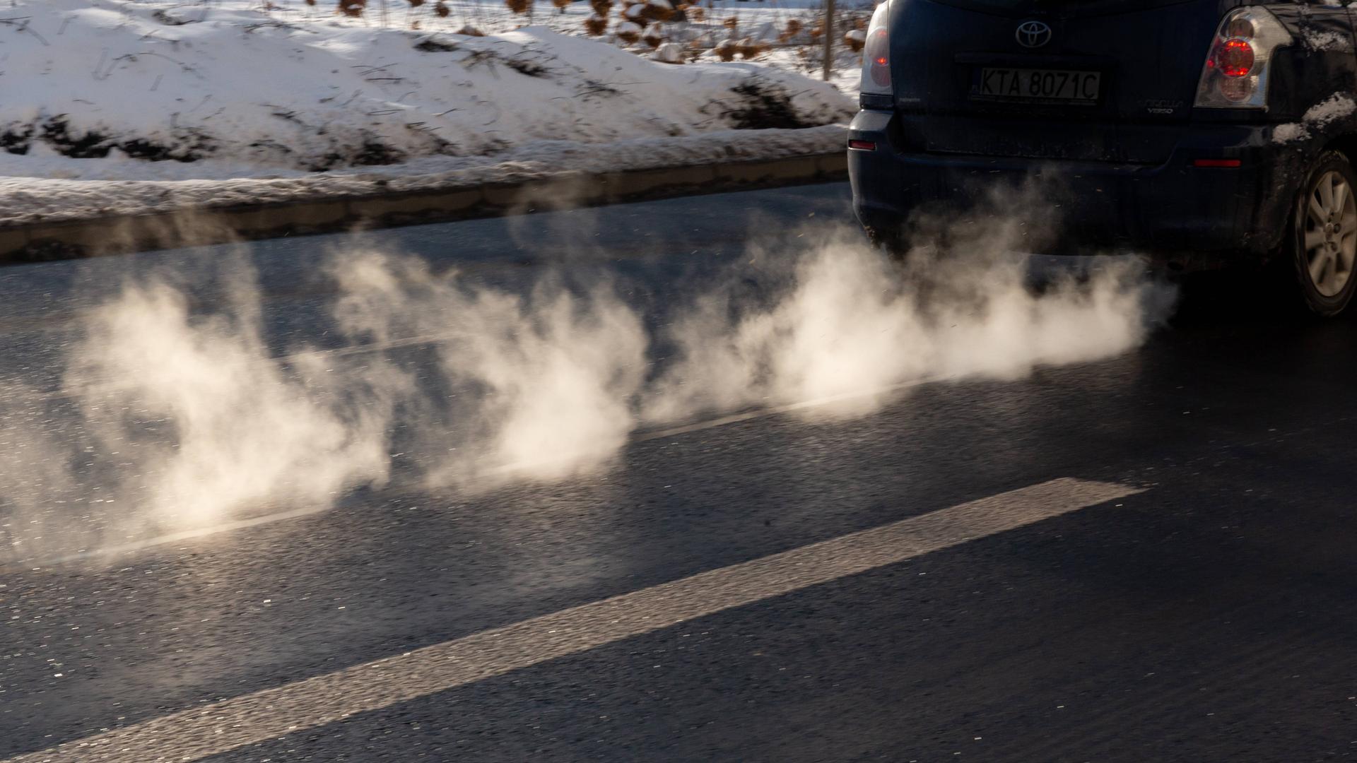 Sichtbare Abgase entweichen aus einem Autoauspuff. Neben der Straße liegt Schnee.