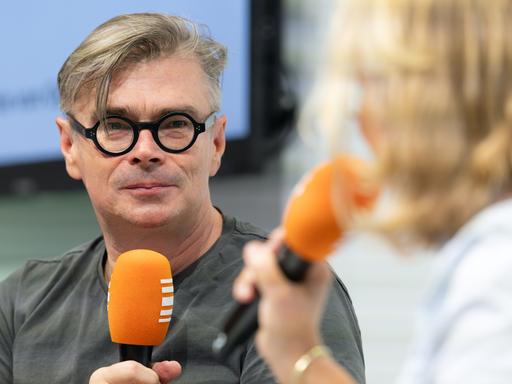 Jaroslav Rudiš trägt eine schwarze umrandete Brille und hält ein Mikrofon in der Hand. Ihm gegenüber sitzt mit Stephanie von Oppen, die ebenfalls ein Mikrofon in der Hand hält.