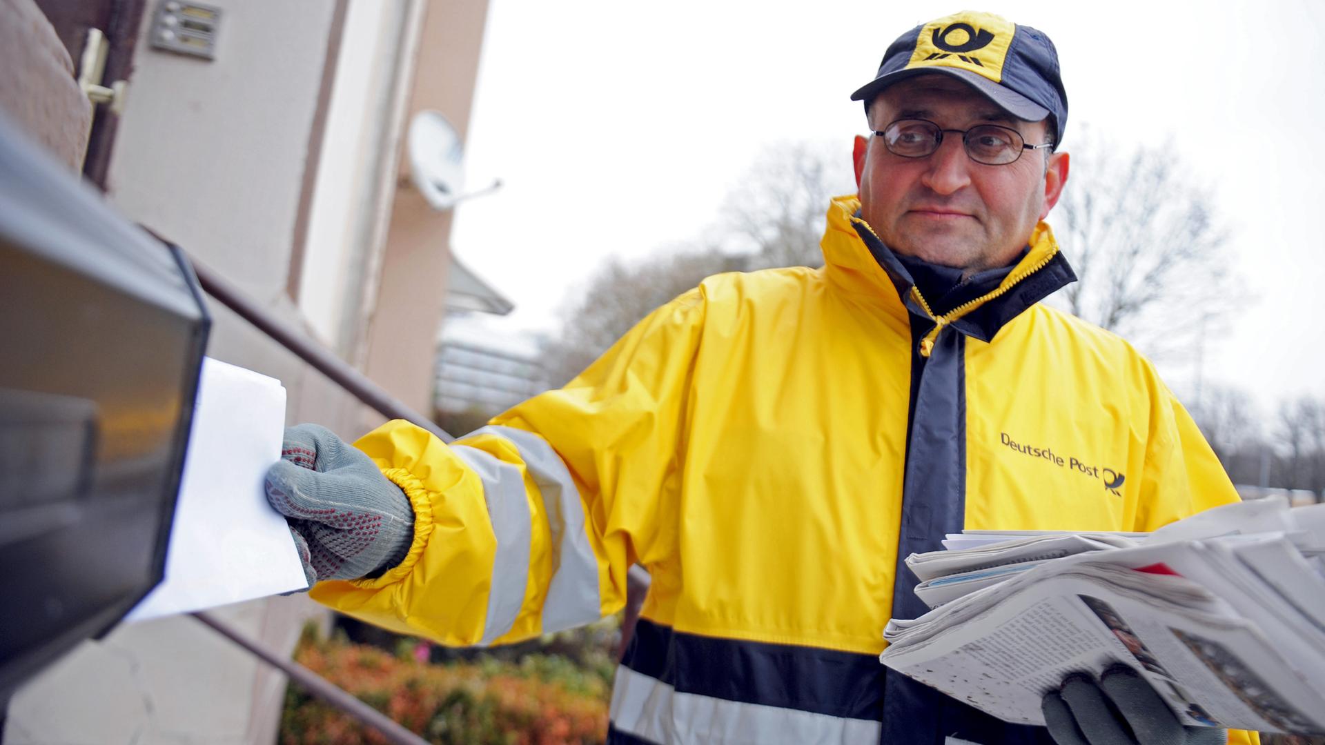 Ein Mann trägt eine Kappe und eine Jacke der Deutschen Post. Er steckt einen Brief in einen Briefkasten an einem Haus. In der Hand hält er weitere Briefe und Zeitungen. 