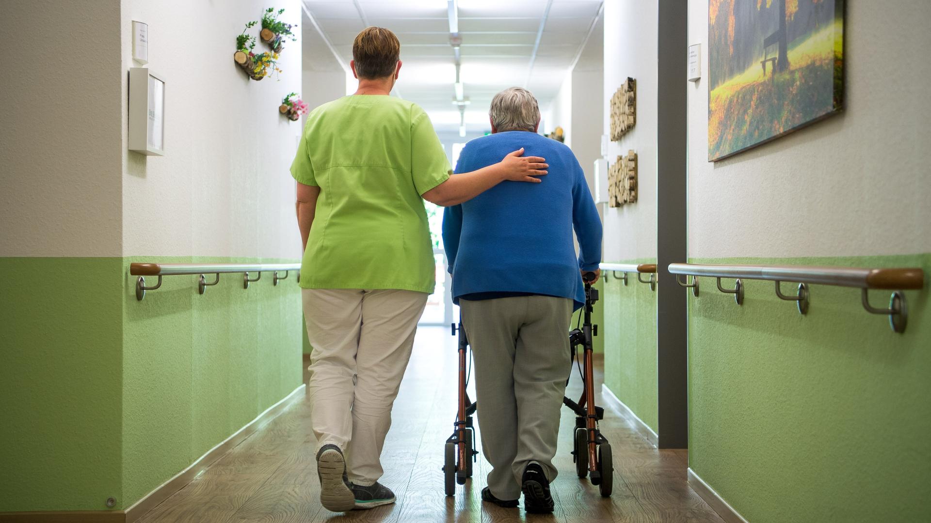 Eine Pflegerin hilft einer alten Frau am Rollator. Sie stützt sie beim Gehen. Beide sind von hinten abgebildet. Eine Pflegerin hilft einer alten Frau am Rollator. Sie stützt sie beim Gehen. Beide sind von hinten abgebildet.