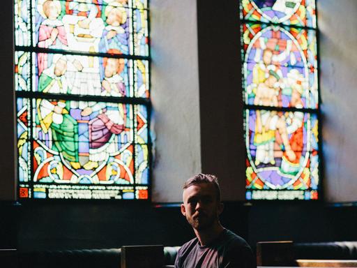 Ein junger Mann sitzt einsam in einer dunklen Kirchenbank, seitlich fällt buntes Licht durch Kirchenfenster in den Raum.