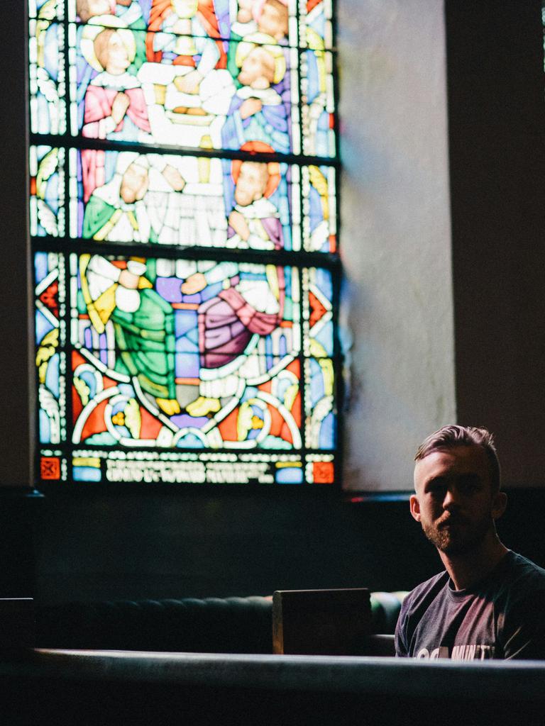 Ein junger Mann sitzt einsam in einer dunklen Kirchenbank, seitlich fällt buntes Licht durch Kirchenfenster in den Raum.