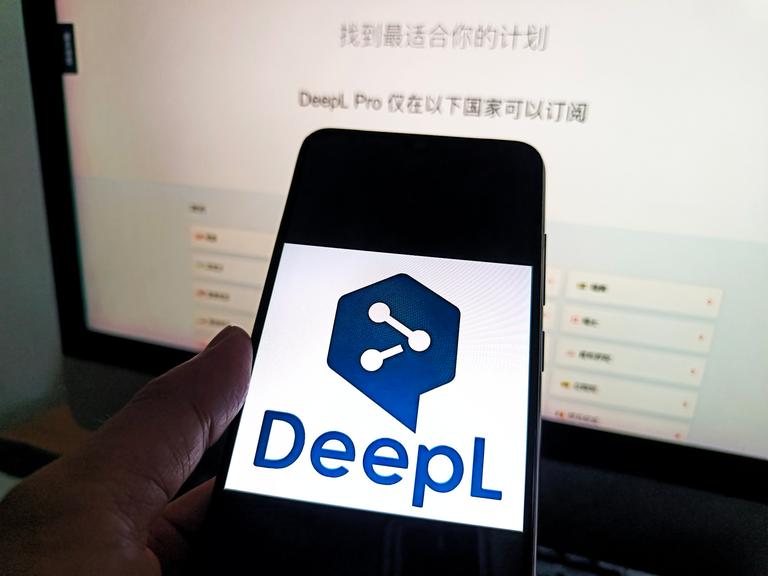 Auf einem Smartphone ist das Logo von DeepL zu sehen, dahinter ist ein Bildschirm.