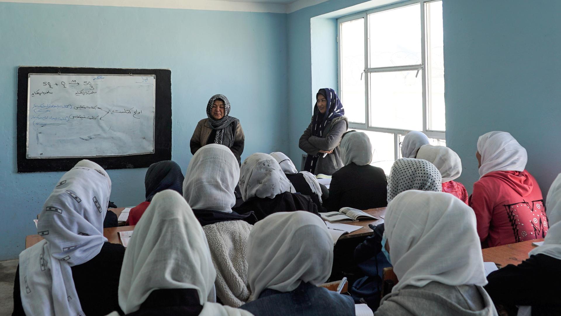 Schülerinnen mit Kopftüchern sitzen in einem Klassensaal; vorne sind zwei Lehrerinnen und ein beschriebenes Whiteboard zu sehen.
