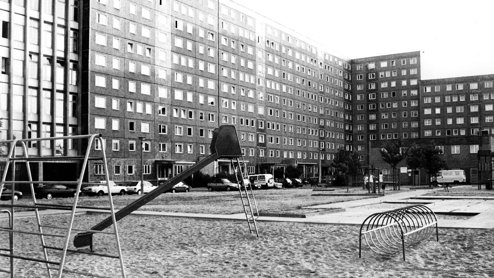 Schwarz-weiß-Foto eines Spielplatzes mit Rutsche und Klettergerüst, der von Hochhäusern in Plattenbauweise umgeben ist