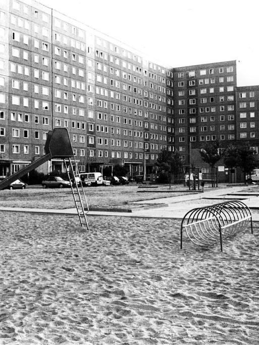 Schwarz-weiß-Foto eines Spielplatzes mit Rutsche und Klettergerüst, der von Hochhäusern in Plattenbauweise umgeben ist