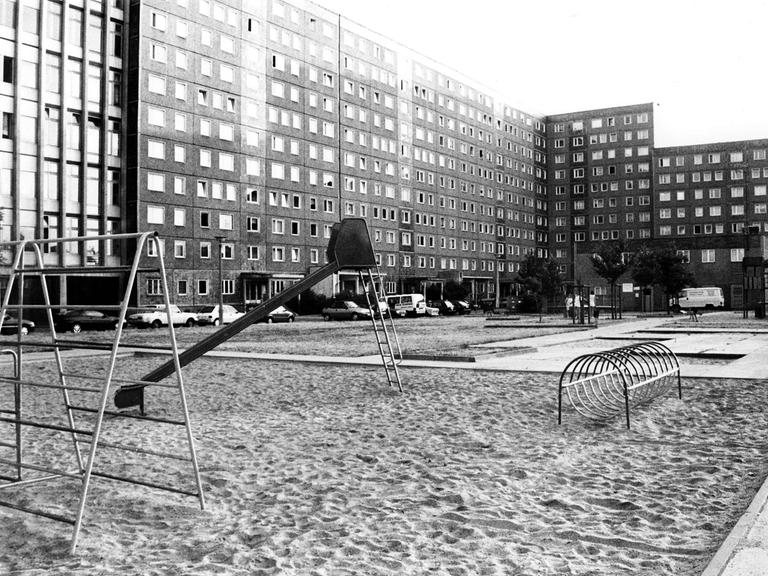 Schwarz-weiß-Foto eines Spielplatzes mit Rutsche und Klettergerüst, der von Hochhäusern in Plattenbauweise umgeben ist