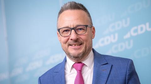 Heiko Strohmann steht vor einem hellblauen Hintergrund mit weißem CDU-Logo, trägt einen blauen Anzug, pinkfarbene Krawatte und eine Brille. Er lächelt direkt in die Kamera.